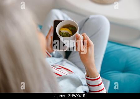 Femme buvant du thé à base de plantes et utilisant un téléphone cellulaire à la maison Banque D'Images