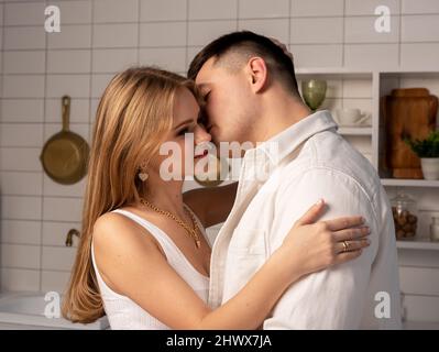 Beau homme embrassant la femme dans la joue dans la cuisine. Un jeune couple heureux qui s'embrasse tout en cuisinant à la maison. Photo de haute qualité Banque D'Images