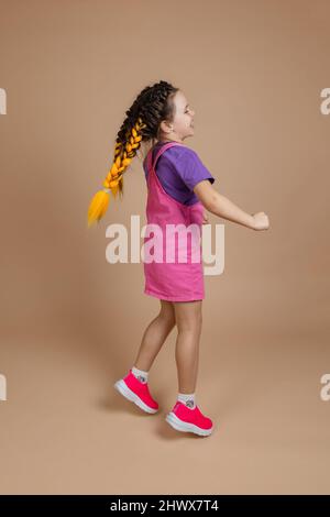 Petite fille dansante gaie avec deux queues de cochon jaune de kanekalon qui sautent en regardant loin en souriant dans une combinaison rose, un t-shirt violet et des baskets roses Banque D'Images