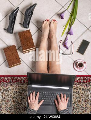 Une femme travaille sur un ordinateur portable assis sur une moquette après avoir pris ses chaussures avec des talons, avec des livres, une tasse de café, un smartphone et des fleurs sur le sol Banque D'Images