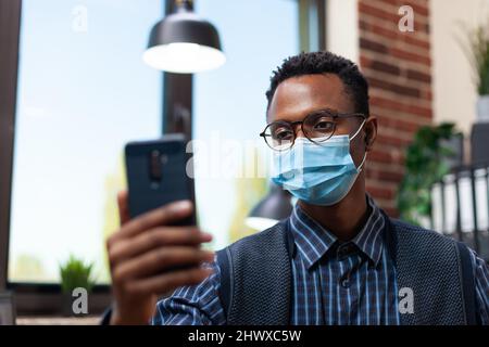 Portrait d'un employé de bureau de démarrage avec des lunettes portant un masque de visage tenant un smartphone dans une interview en ligne. Concentrez-vous sur l'entrepreneur en isolement dans les appels vidéo regardant l'écran de téléphone mobile. Banque D'Images