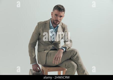 homme en costume avec bagages assis sur une chaise en bois et prenant quelques jours de congé pour partir en vacances, posant sur fond gris Banque D'Images