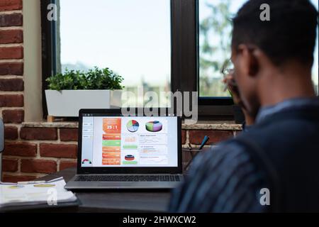 Vue d'ensemble de l'employé d'une start-up afro-américaine qui regarde l'écran d'un ordinateur portable avec des tableaux d'analyse d'entreprise assis à son bureau. Gros plan sur l'affichage des ordinateurs portables avec les résultats des ventes. Banque D'Images