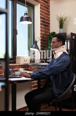 Entrepreneur blessé avec col cervical de retour au travail après un traumatisme physique au bureau de démarrage. Afro-américain assis portant un corset de cou se rétablissant après un accident de travail sur un ordinateur portable. Banque D'Images