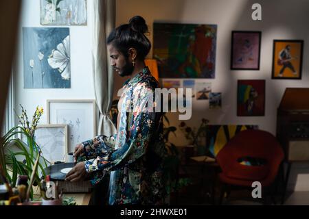 Jeune homme contemporain d'origine indienne mettant le disque de vinyle dans le joueur tout en allant écouter de la musique à la maison Banque D'Images