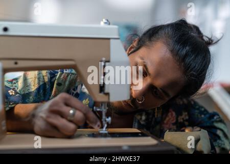 Un homme sérieux de l'ethnie indienne mettant le fil dans l'oeil d'aiguille de la machine à coudre électrique tout en travaillant sur la nouvelle collection de mode Banque D'Images