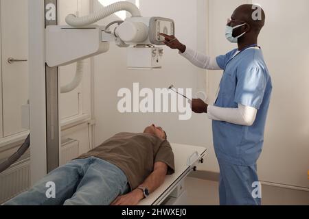 Jeune clinicien en uniforme et masque de protection allumer la machine à rayons X avant d'examiner un patient mâle mature à l'hôpital Banque D'Images