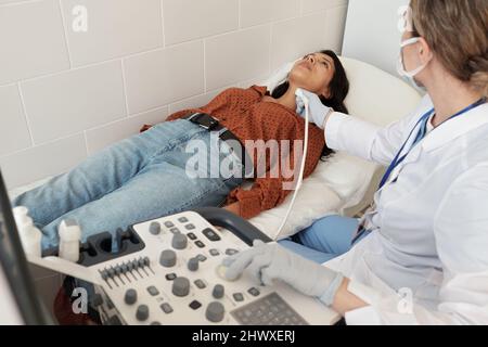 Jeune médecin en uniforme et gants examinant la thyroïde de la patiente avec dispositif d'échographie dans le bureau médical des cliniques modernes Banque D'Images