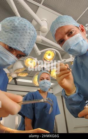 Une équipe chirurgicale de trois médecins – deux hommes et une femme – portant des robes, des casquettes et des masques bleus, à l'intérieur d'un centre d'opération hospitalier sous les frais généraux Banque D'Images