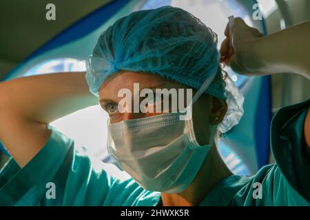 Une femme chirurgien attache son masque chirurgical autour de son visage sous les lumières du théâtre (MODÈLE RELÂCHÉ) Banque D'Images