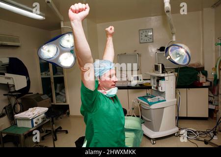 Un chirurgien fatigué surpassé étire ses bras pendant qu'il est assis sur une table d'opération. (MODÈLE DISPONIBLE) Banque D'Images