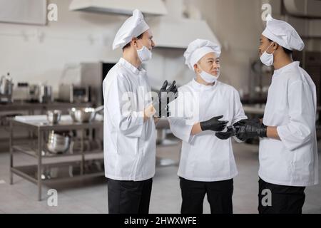 Trois chefs multiraciaux ont une conversation dans la cuisine professionnelle. Un chef asiatique s'entretient avec des cuisiniers latino et européens lors d'une pause au restaurant. Des chefs bien habillés en gants de protection et masques de protection au travail Banque D'Images