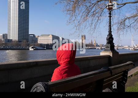 Une personne portant un sweat à capuche rouge s'assoit sur un banc au bord de la rivière qui domine la Tamise où MI5 le service de renseignements du Royaume-Uni est situé, le 7th mars 2022, à Londres, en Angleterre. Banque D'Images