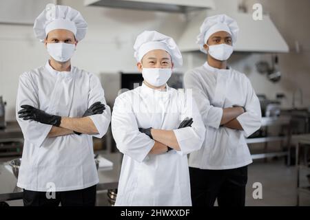 Portrait d'une équipe multiraciale composée de trois chefs réunis dans la cuisine professionnelle. Chefs bien habillés, masques faciaux et gants de protection prêts pour un travail. Nouvelle norme pour les affaires en cas de pandémie Banque D'Images