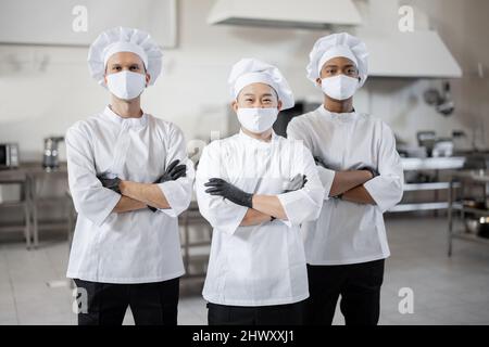 Portrait d'une équipe multiraciale composée de trois chefs réunis dans la cuisine professionnelle. Chefs bien habillés, masques faciaux et gants de protection prêts pour un travail. Nouvelle norme pour les affaires en cas de pandémie Banque D'Images