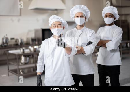 Portrait d'une équipe multiraciale composée de trois chefs réunis dans la cuisine professionnelle. Chefs bien habillés, masques faciaux et gants de protection prêts pour un travail. Nouvelle norme pour les affaires en cas de pandémie Banque D'Images