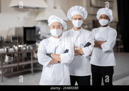 Portrait d'une équipe multiraciale composée de trois chefs réunis dans la cuisine professionnelle. Chefs bien habillés, masques faciaux et gants de protection prêts pour un travail. Nouvelle norme pour les affaires en cas de pandémie Banque D'Images