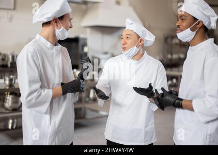 Trois chefs multiraciaux ont une conversation dans la cuisine professionnelle. Un chef asiatique s'entretient avec des cuisiniers latino et européens lors d'une pause au restaurant. Des chefs bien habillés en gants de protection et masques de protection au travail Banque D'Images