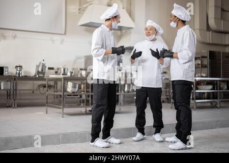 Trois chefs multiraciaux ont une conversation dans la cuisine professionnelle. Un chef asiatique s'entretient avec des cuisiniers latino et européens lors d'une pause au restaurant. Des chefs bien habillés en gants de protection et masques de protection au travail Banque D'Images