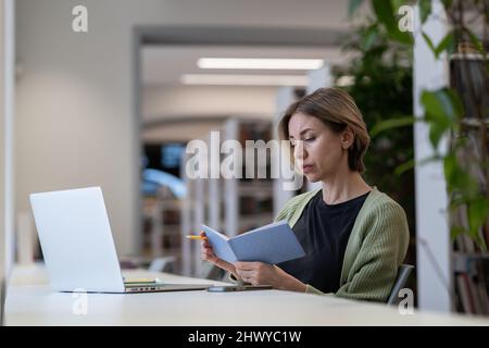 Femme ciblée écrivant une liste de tâches dans un ordinateur portable tout en travaillant à distance sur un ordinateur portable dans une bibliothèque calme et confortable Banque D'Images