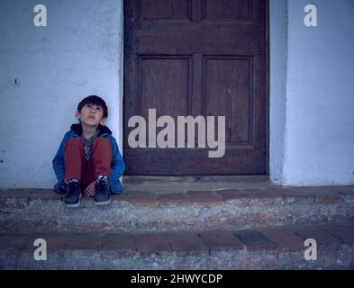 latino petit garçon est assis sur les escaliers près de la porte de la maison donnant vers le haut, attendant pensive et triste. Environnement horizontal et spectaculaire. Banque D'Images