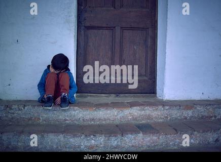 Melancholy Mixed Race Boy assis à l'extérieur de sa maison en colère et triste avec sa tête reposant sur ses jambes. Horizontal, latino enfant regardant vers le bas Banque D'Images