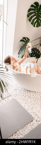 Gaie jeune femme s'amusant en versant du gel douche sur l'éponge loofah, en prenant le bain dans la salle de bains moderne décorée de plantes tropicales Banque D'Images