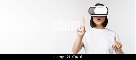 Potrait de la femme asiatique dans les lunettes de réalité virtuelle, pointant, choisissant smth dans le casque VR, debout sur fond blanc Banque D'Images