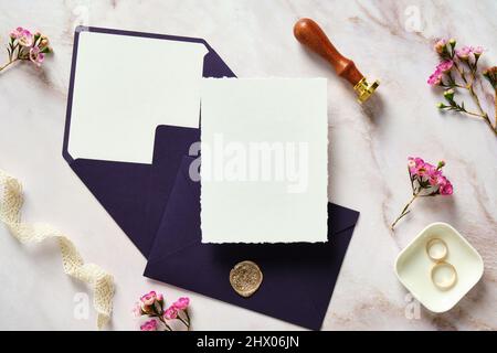 Élégante papeterie de mariage. Modèle de carte d'invitation de mariage, enveloppes violettes, ruban en soie, anneaux, fleurs roses sur table de bureau en pierre. Pose à plat, à Banque D'Images