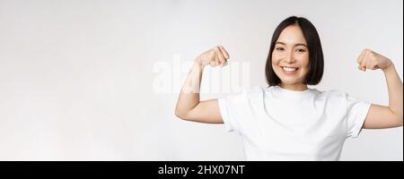 Femme asiatique souriante montrant des biceps flexibles, des muscles forts bras gestuelle, debout dans un t-shirt blanc sur fond blanc Banque D'Images