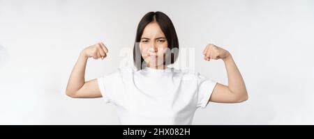 Femme asiatique souriante montrant des biceps flexibles, des muscles forts bras gestuelle, debout dans un t-shirt blanc sur fond blanc Banque D'Images