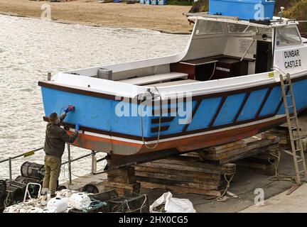 New Quay, pays de Galles - Mars 2022: Personne utilisant une ponceuse électrique pour préparer la coque d'un bateau à moteur pour la peinture prête pour la saison touristique d'été Banque D'Images