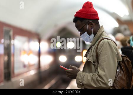 Homme africain dans le masque de visage dans la station de métro attendre le train, en utilisant le smartphone, défilement des médias sociaux Banque D'Images