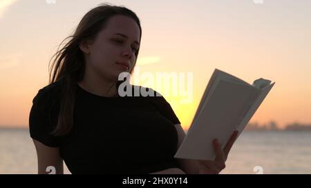 Une jeune femme lit un livre en étant couché sur la plage au coucher du soleil Banque D'Images