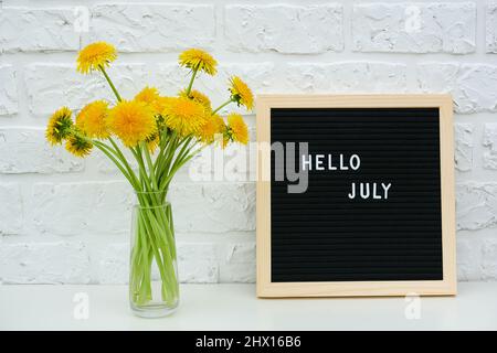 Texte Bonjour juillet sur le tableau noir de lettres et le bouquet de fleurs de pissenlits jaunes dans un vase sur fond blanc mur de brique. Concept de modèle de carte de vœux. Banque D'Images