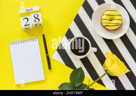 Cubes en bois calendrier avril 28th. Tasse de café, beignet jaune et rose sur serviette noire et blanche, bloc-notes vide ouvert pour le texte sur fond jaune. Co Banque D'Images