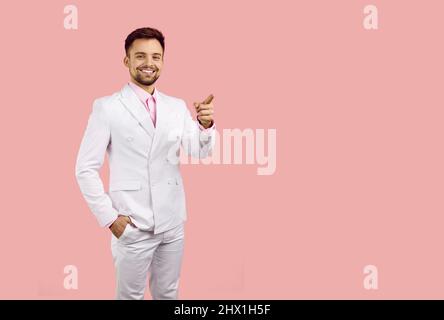Homme souriant en costume blanc vous regarde et pointe son index vers l'espace de copie sur fond rose. Banque D'Images