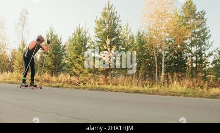 Entraînement d'un athlète sur les patineurs à roulettes. Biathlon sur les skis à roulettes avec des bâtons de ski, dans le casque. Entraînement d'automne. Rouleau sport. Homme adulte sur patins. Tirer un athlète en mouvement. Stabilisateur de plate-forme mobile. Ralenti. Banque D'Images