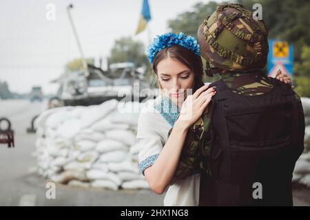 Le soldat ukrainien embrasse une jeune femme en robe nationale traditionnelle sur la barrière contre le fond des sacs de sable. Concept d'invasion militaire russe Banque D'Images