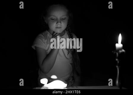 Une petite fille dessine des personnages de fantaisie près d'une bougie dans une pièce sombre. Enfant faisant ce qu'il préfère lors d'une panne de courant. En noir et blanc. Banque D'Images