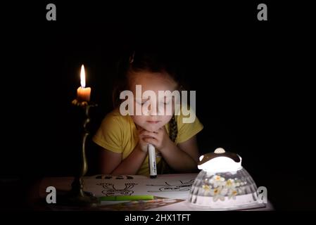 Une petite fille dessine des personnages de fantaisie près d'une bougie dans une pièce sombre. Enfant faisant ce qu'il préfère lors d'une panne de courant. Banque D'Images