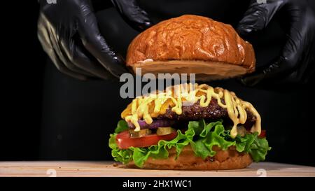 Le hamburger artisanal est en cours de cuisson sur fond noir avec des gants alimentaires noirs. Se composent de sauce, laitue, tomate, oignon rouge, cornichon, fromage, bacon, pain à l'air et viande de bœuf marbré. Pas idéal. Il a l'air réel, affectueux fait main. Banque D'Images
