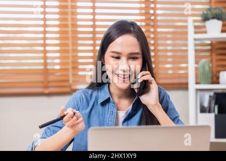 Une femme d'affaires parle au téléphone et écrit sur un ordinateur portable, une fille asiatique travaille à la maison Banque D'Images