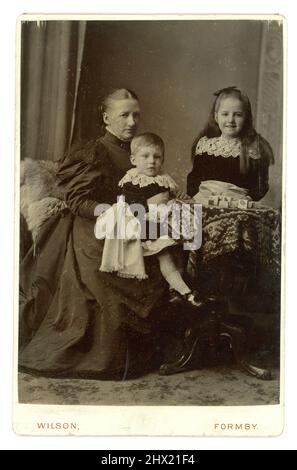 Charmant original victorien carte de cabinet portrait studio de mère de classe moyenne avec sa fille souriante, et jeune fils - les deux enfants sont vêtus de velours avec des cols de dentelle blanche (style Little Lord Fauntleroy). La mère porte une robe tendance avec de grandes manches bouffantes (connue sous le nom de jambe de manchons de mouton). Typique de la mode en 1894. La photographie est datée de mars 16th 1894 du studio photographique M. W. Wilson, Formby, Angleterre, Royaume-Uni Banque D'Images