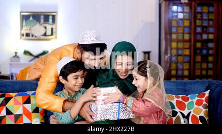 Un beau gars avec une barbe apportant des cadeaux colorés pour ses enfants à la maison. Une mère musulmane portant un hijab et ses deux jeunes enfants assis ensemble Banque D'Images