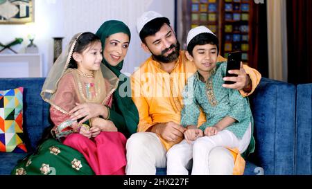 Une famille musulmane joyeuse effectuant un appel vidéo sur son smartphone à la maison. Une famille indienne joyeuse utilisant un mobile ensemble tout en étant assis ensemble Banque D'Images