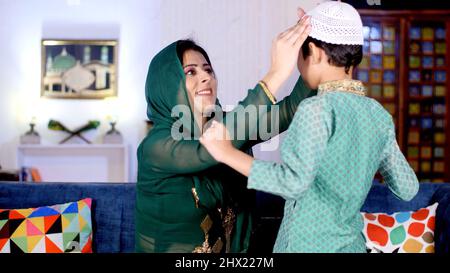 Une femme indienne heureuse portant un foulard fait porter un chapeau à son fils. Un enfant musulman dans une robe ethnique et sa belle mère ensemble dans le salon Banque D'Images