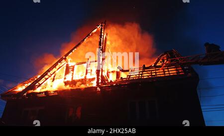 Le toit de la maison est en feu. Le bâtiment résidentiel brûler, village. Les pompiers ont tiré un incendie de la grue par le dessus. Le service des incendies qui produit la flamme nue. Fumée et étincelles. Banque D'Images