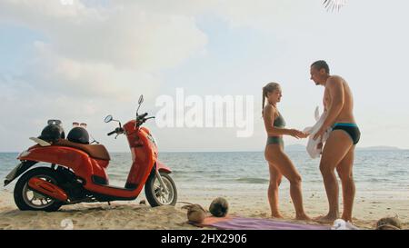 Trajet en scooter. Un couple charmant sur une moto rouge vêque de vêtements blancs sur une plage de sable. Les gens juste mariés embrassent les hugs marchant près des palmiers tropicaux, la mer. Lune de miel de mariage au bord de l'océan. Location de motos. Banque D'Images