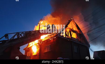 Le toit de la maison est en feu. Le bâtiment résidentiel brûler, village. Les pompiers ont tiré un incendie de la grue par le dessus. Le service des incendies qui produit la flamme nue. Fumée et étincelles. Banque D'Images